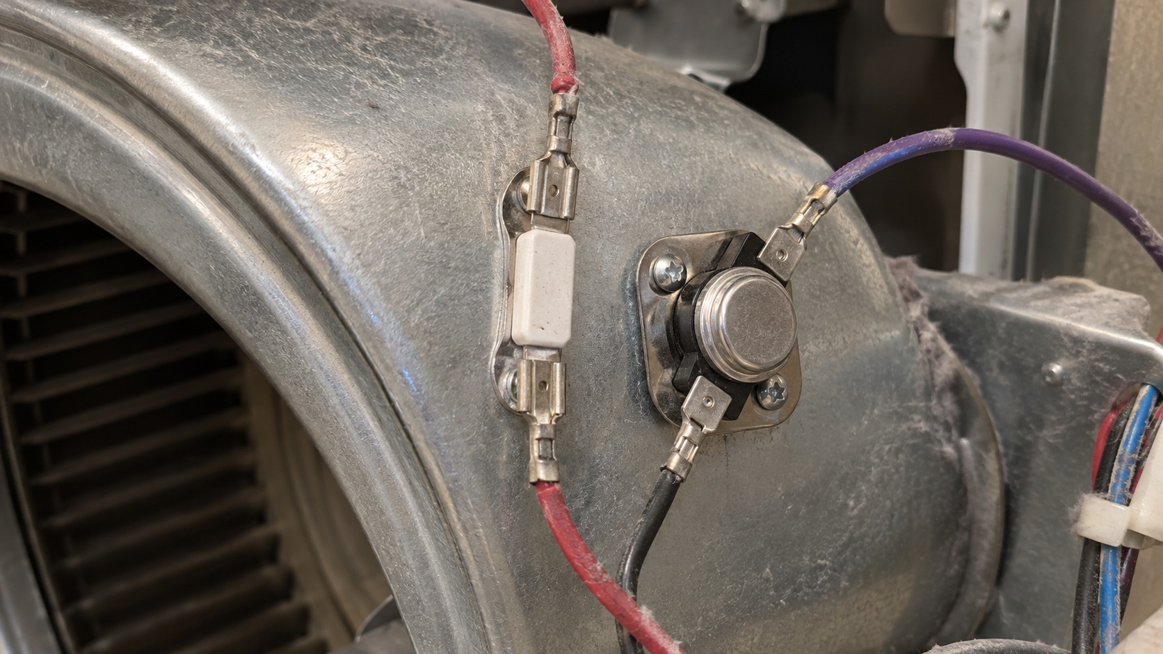 Macro close-up of a white ceramic thermal fuse and a round metal cycling thermostat mounted on the sheet-metal blower housing inside a clothes dryer.