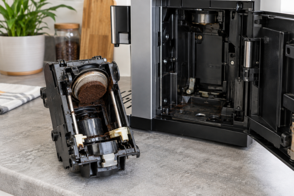A brewing unit taken out of a coffee machine and set on the counter beside the open service door, with a pressed coffee puck stuck to its screen.