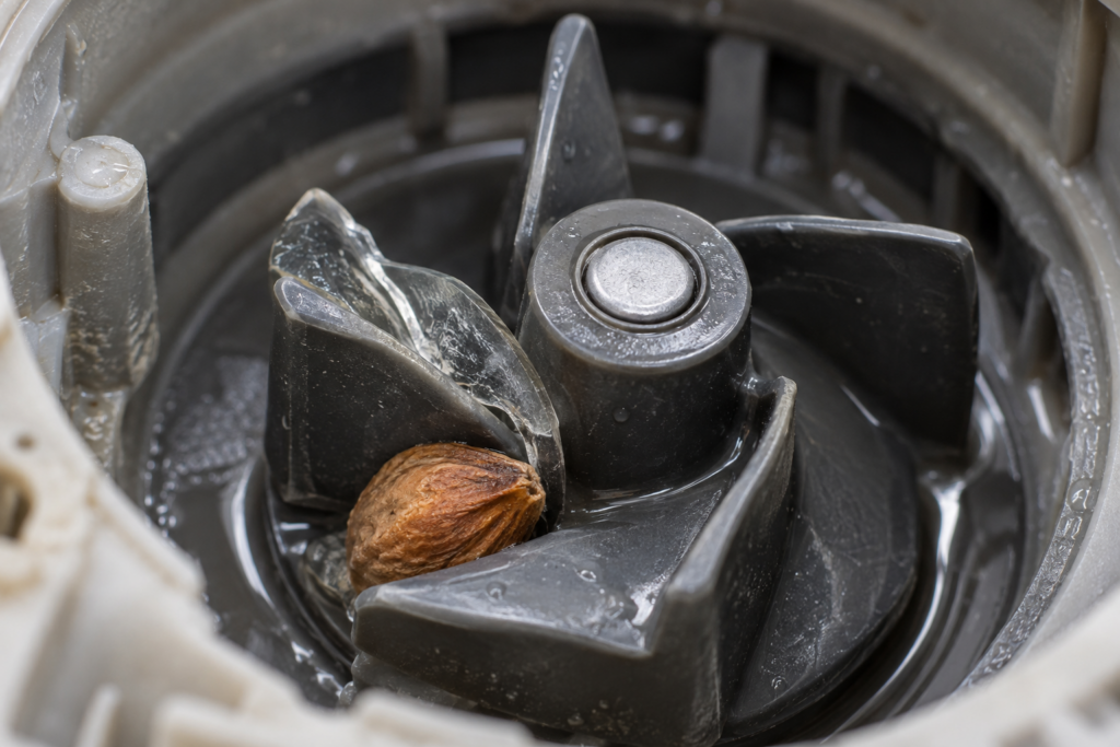 A macro view of a dishwasher drain pump impeller with a small curved glass shard wedged between two blades and a fruit pit beside it, even side light, blurred sump housing behind.