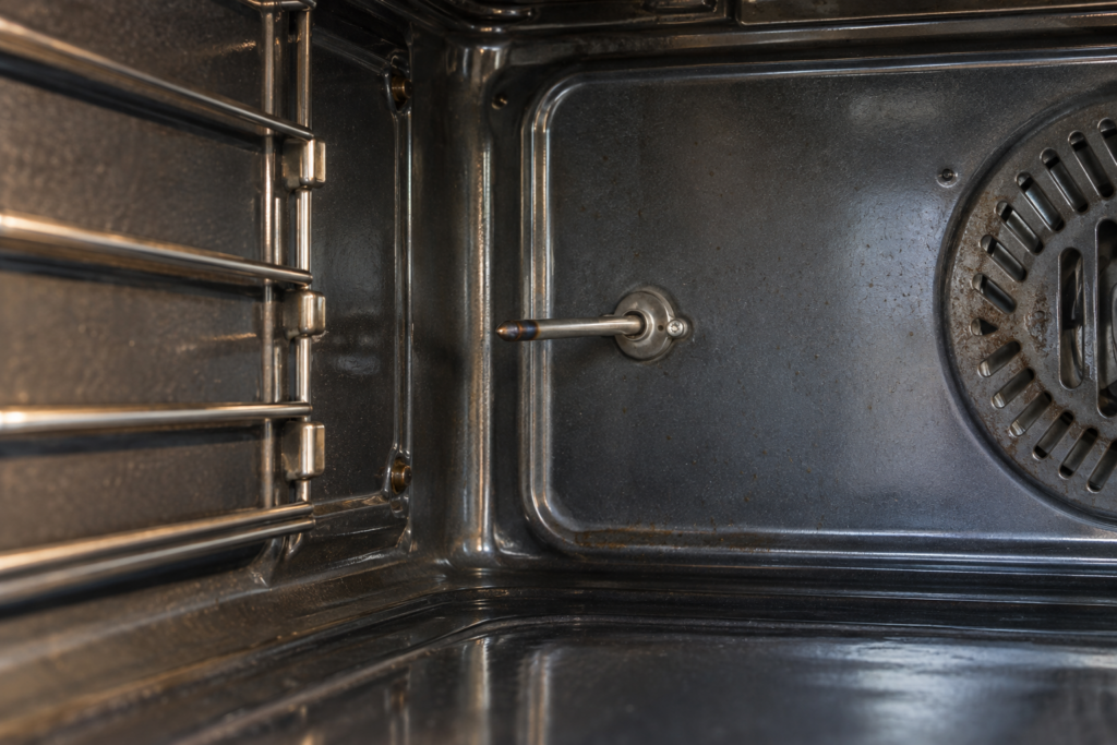 A slim metal oven temperature probe projecting from the rear wall of an empty oven cavity, seen from the open door at a low angle with its tip slightly darkened, even light.
